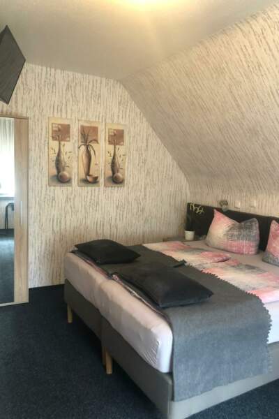 Gemütliches Doppelzimmer mit großem Bett, grauen und rosafarbenen Kissen und Decken, Wandbildern sowie einem Spiegel am Schrank