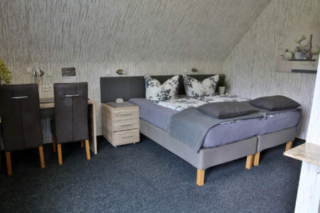 Hotelzimmer mit Doppelbett, grauer Bettwäsche, floral gemusterten Kissen, Schreibtisch mit zwei Stühlen und Nachttisch.